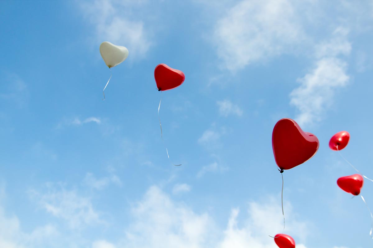 Red and white heart balloons float into a blue sky with light clouds.